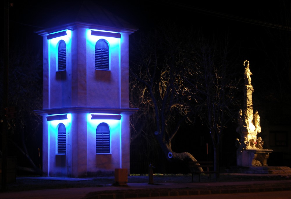 Obr. 48. Iluminácia mestskej zvonice v Hurbanove modrým svetlom žiariviek v kontraste s teplým bielym osvetlením sochy sv. Jána Nepomuka vpravo; v súčasnosti je budova zvonice nadstavaná o jedno podlažie a má novú ilumináciu